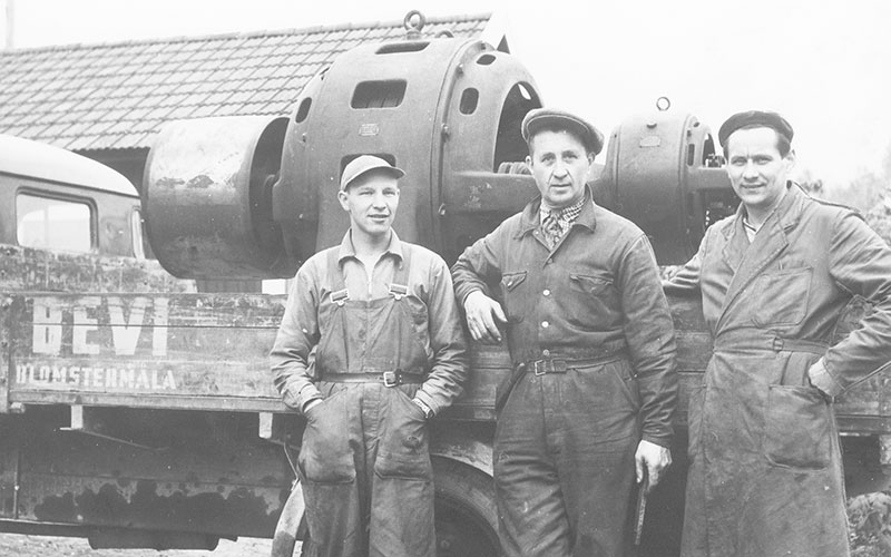 Tre mænd i arbejdstøj ved en lastbil med en stor elmotor på flatbed, omkring 1940.
